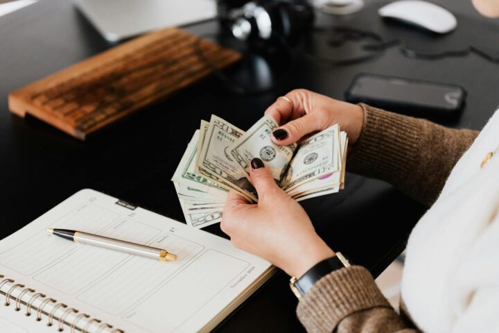 Femme élégante méconnaissable en pull comptant des billets de dollars tout en étant assise à une table en bois avec un agenda et un stylo